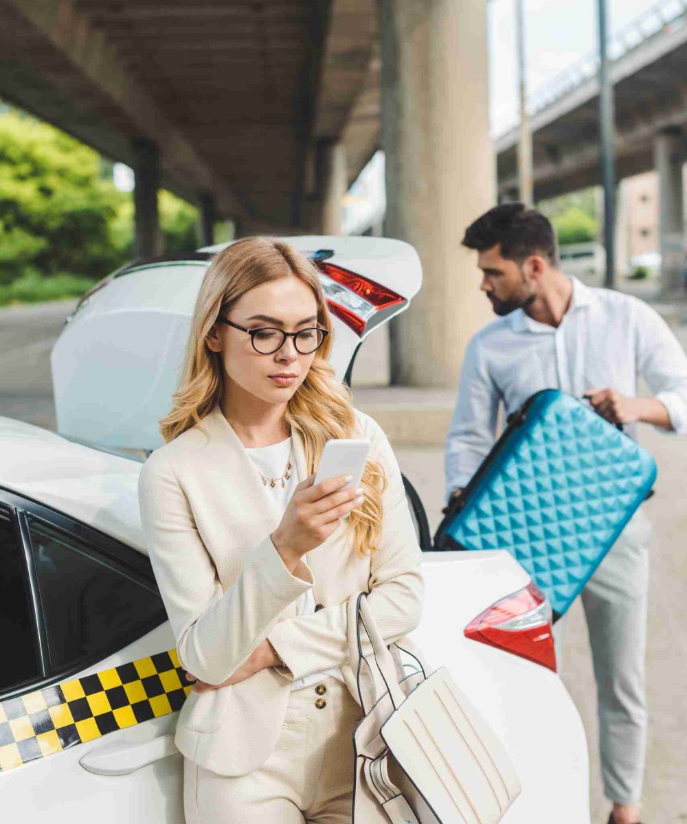 young blonde woman in eyeglasses using smartphone while man putting suitcase in trunk of taxi car